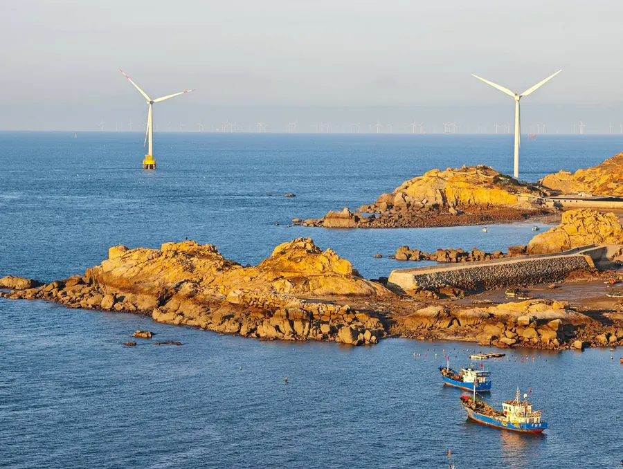 Offshore wind turbines beyond a rocky coastline at low sun, fishing boats moored inshore
